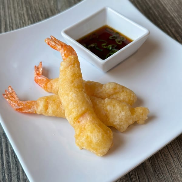 Three golden-brown tempura shrimp with tails on, served on a white plate with a side of dipping sauce.