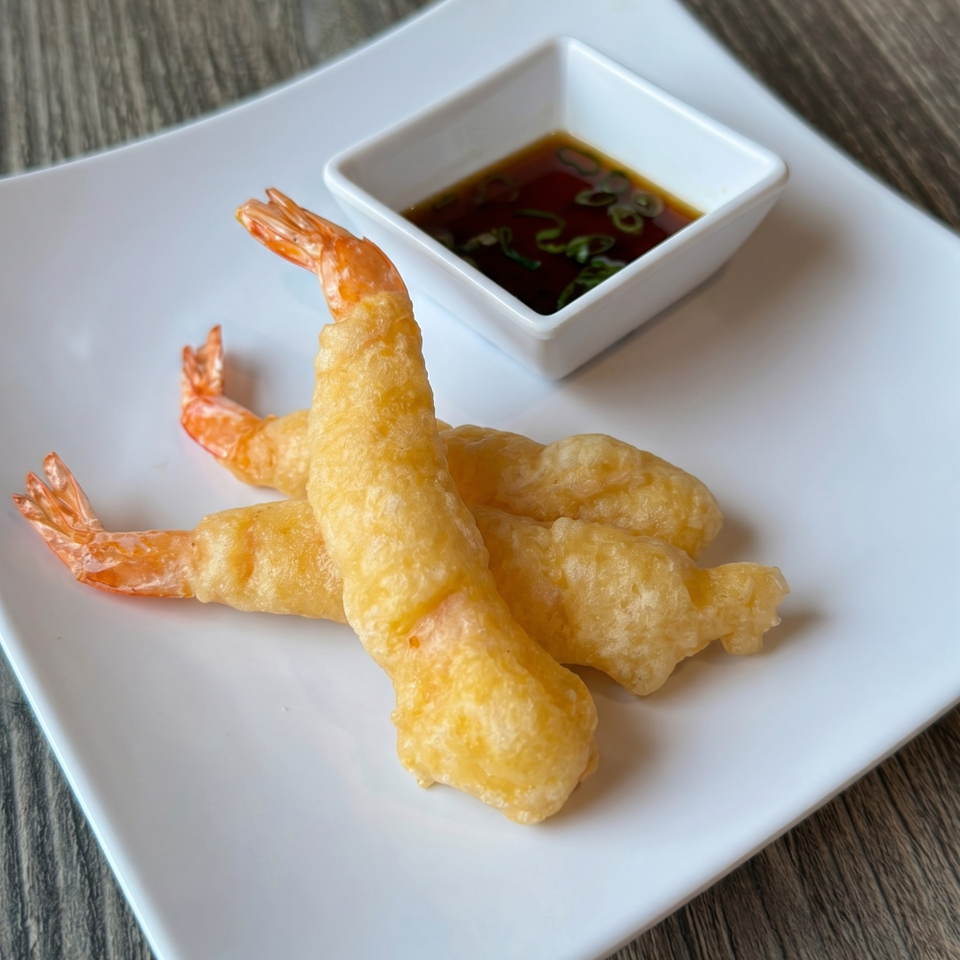Three golden-brown tempura shrimp with tails on, served on a white plate with a side of dipping sauce.
