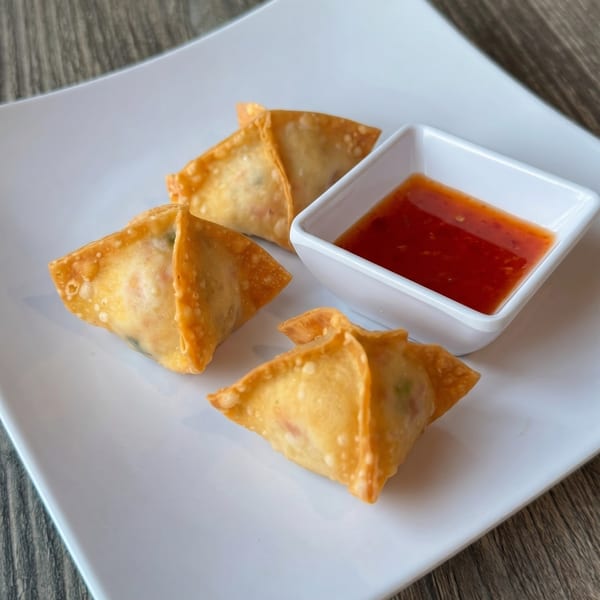 Three golden-fried crab rangoon appetizers with a side of sweet and sour dipping sauce on a white plate.