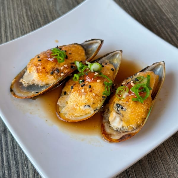 Three baked mussels in shells with orange sauce, sesame seeds, and green onions on a white plate.